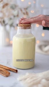 Una mano coloca la tapa en un frasco de cristal Vegan Milker lleno de una bebida cremosa amarillenta; cerca hay palitos de canela.