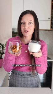 Mujer con jersey rosa usando Vegan Milker y sosteniendo un vaso con almendras y coco rallado en su cocina.