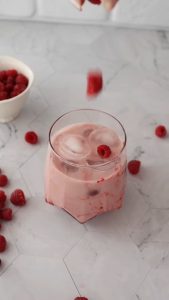 Vaso con bebida helada rosa Vegan Milker y frambuesas, con frambuesas esparcidas sobre una encimera clara.
