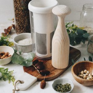 Vegan Milker con base de madera, dátiles, semillas y frutos secos sobre una tabla de madera.