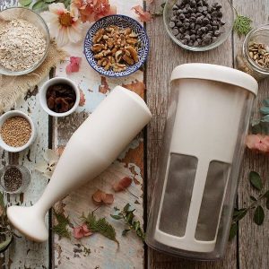 Batidora de inmersión Vegan Milker sobre una mesa rústica, con copos de avena, semillas y virutas de chocolate cerca.