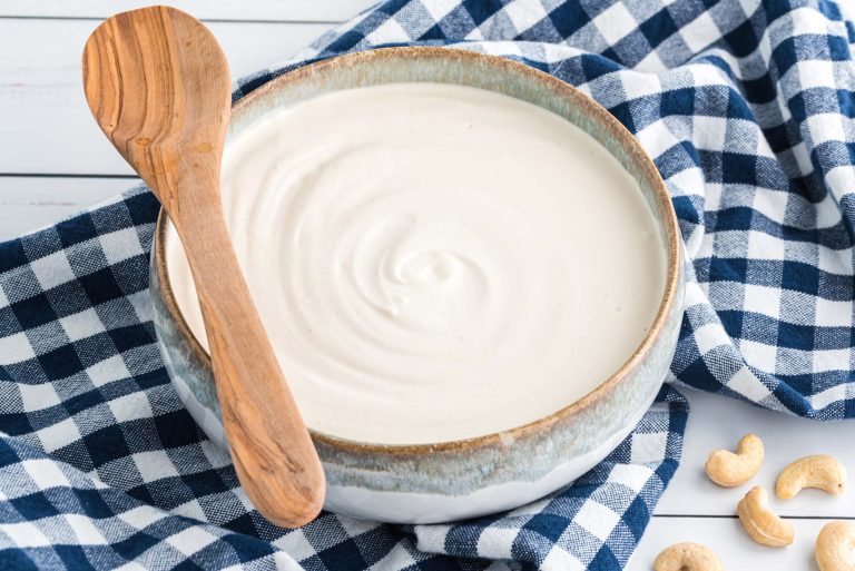 Batidora blanca, tres tarros con tapas metálicas y rojas con una mezcla beige tipo yogur con frutas; nueces, canela y una bolsita de té sobre mesa de madera.