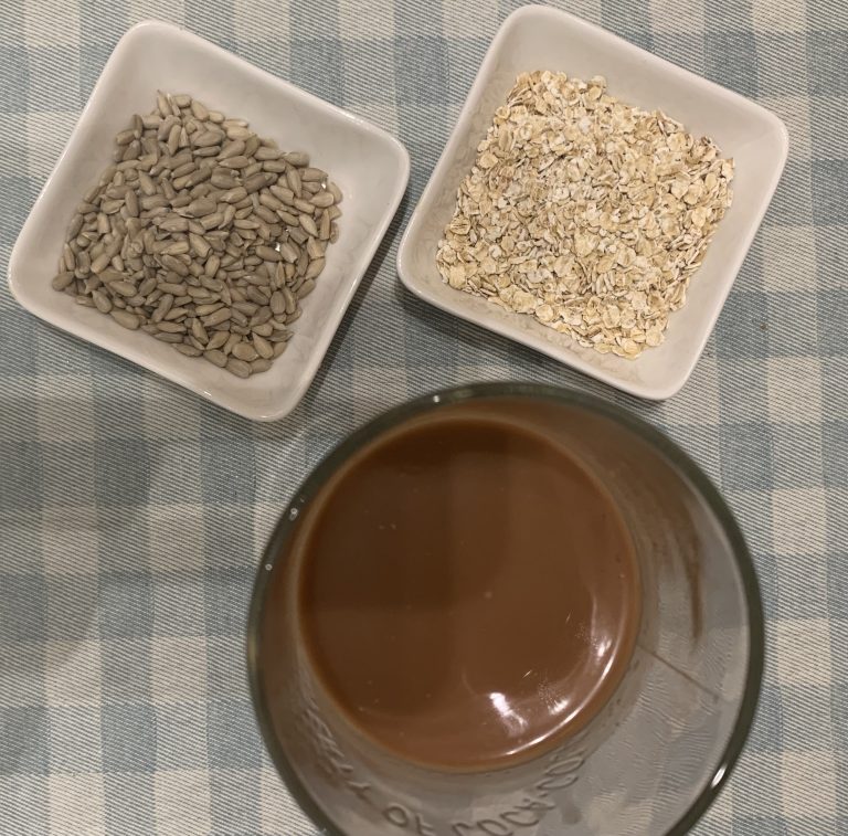 Batido proteico de choclate sobre una mesa junto a dos boles, uno con semillas de girasol y otro con copos de avena.