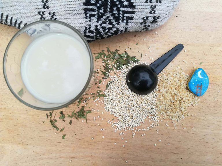 vaso con leche de quinoa sobre mesa de madera junto a copos y granos de quinoa y un gorro con motivos andinos